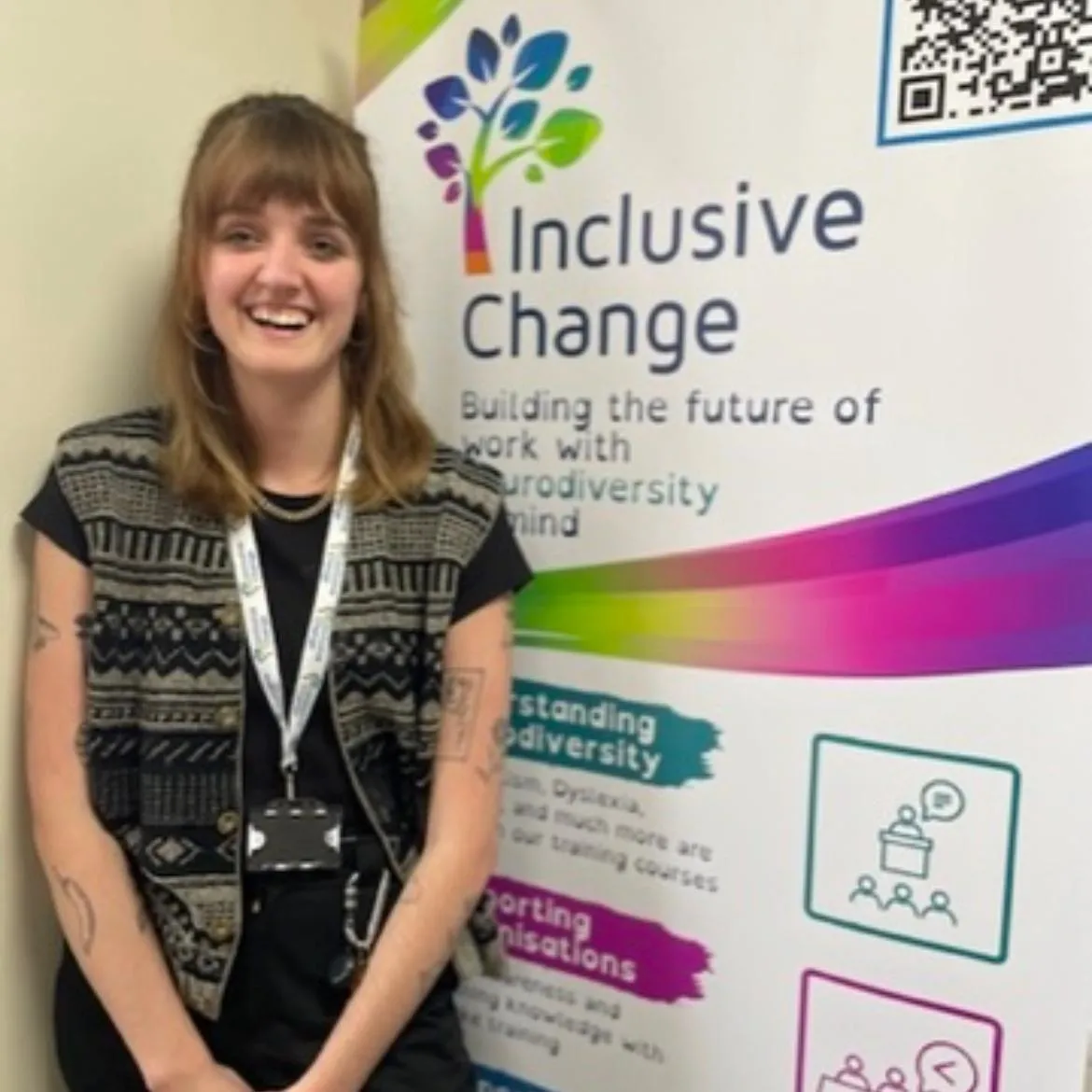 Holly Smith, a smiling young woman with shoulder-length light brown hair, wearing a patterned vest over a black top and a lanyard, and support operations team manager of Inclusive Change, standing in front of an Inclusive Change banner.