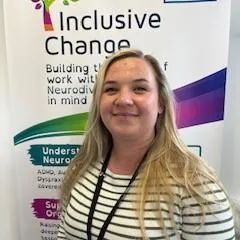 Becca Burke, a smiling woman with long blonde hair, wearing a black-and-white striped top and a lanyard, and operational support specialist of Inclusive Change, standing in front of an Inclusive Change banner.