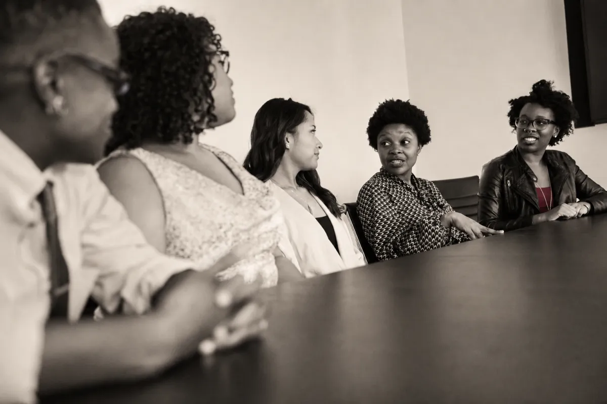 A diverse group of professional women discussing leadership challenges around a conference table in a warm editorial monochrome photograph.