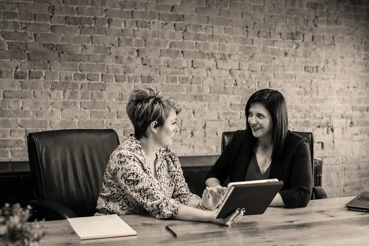 Two experienced professional women reviewing a tablet in a focused meeting, shown in a warm editorial monochrome photograph.