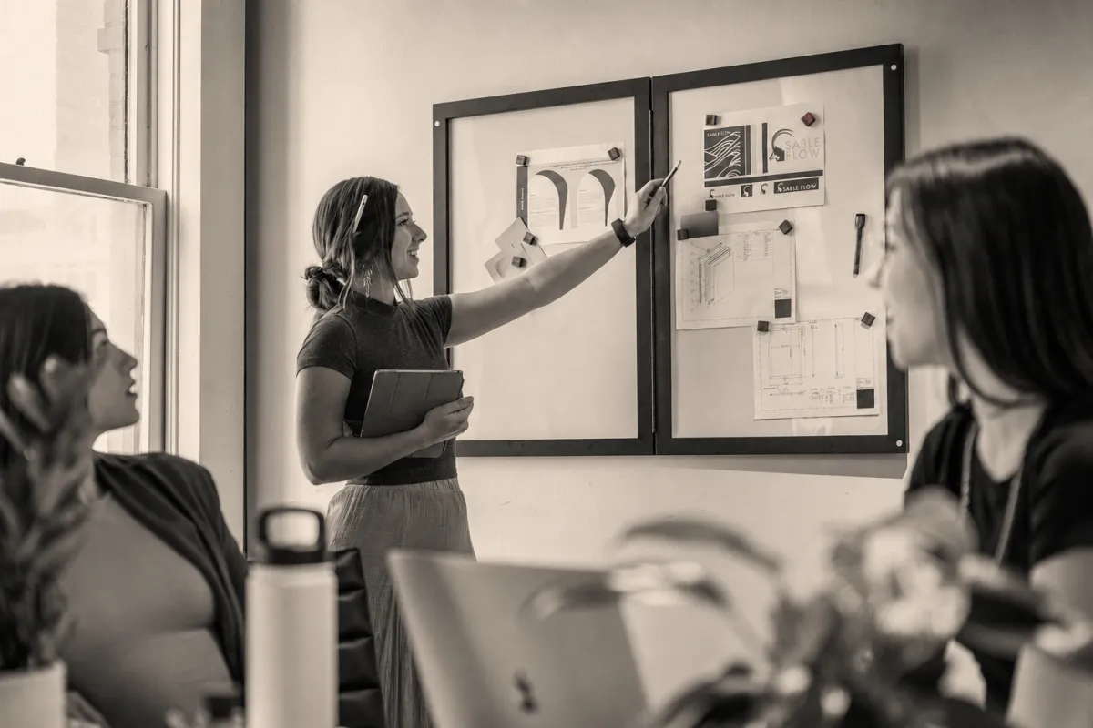 A professional woman aligning colleagues around a shared plan at a strategy board, shown in a warm editorial monochrome photograph.