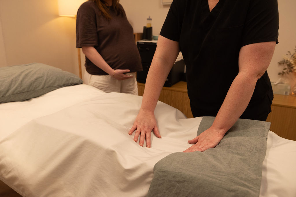 Massage therapist preparing a massage bed while a pregnant woman waits nearby.