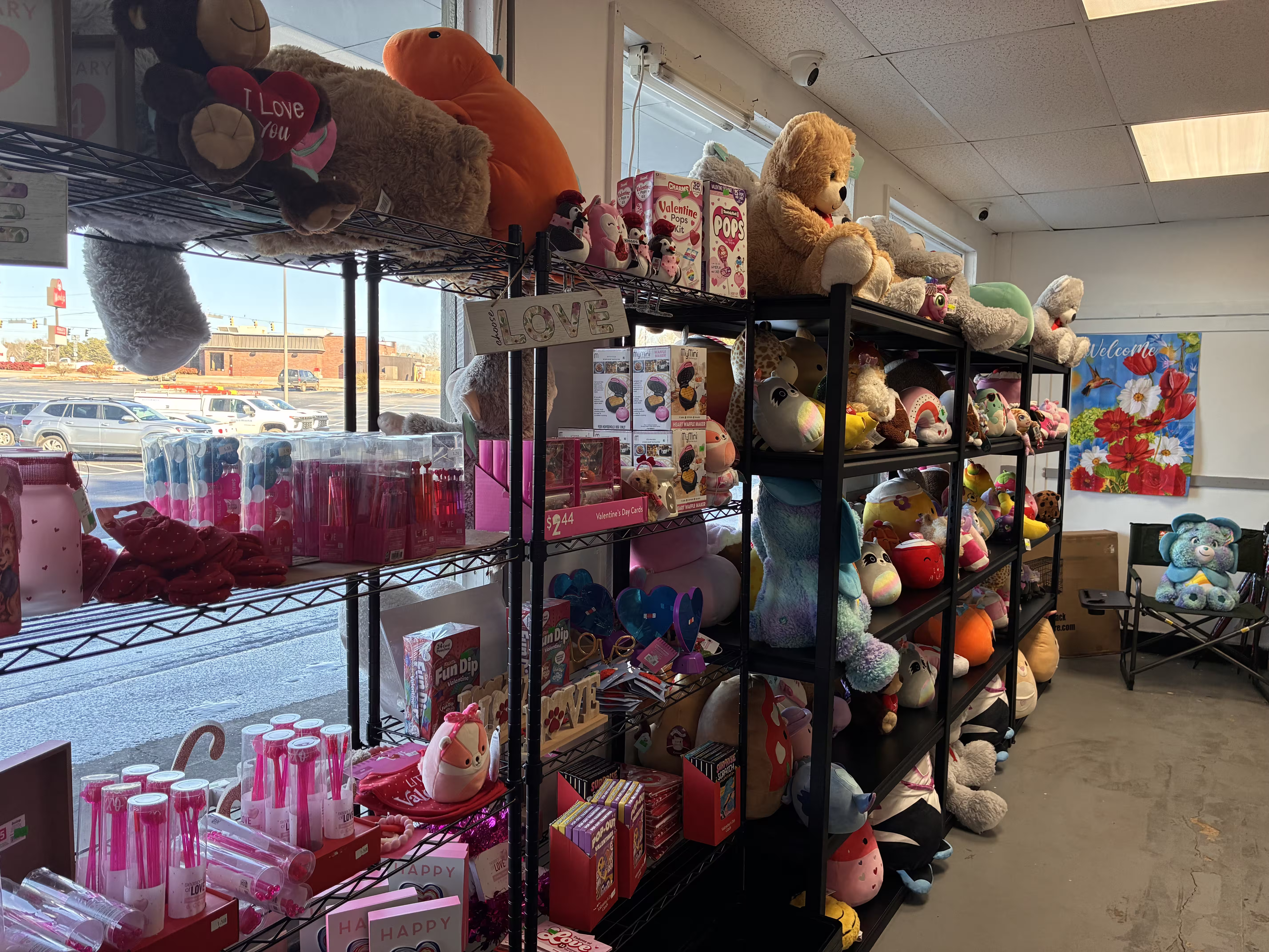 Store aisle with neatly organized shelves displaying folded white and gray bath towels on the top shelf, packaged fleece and plush blankets in soft neutral colors on the middle shelves, and assorted comforters, quilt sets, and bedding bundles on the lower shelves, including patterned and solid-color designs, all arranged in clear plastic packaging under bright retail lighting with a clean walkway visible to the right.