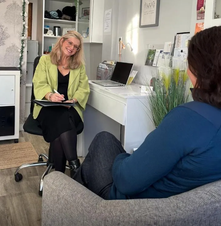 A close-up of a supportive coaching session: Carrie and a client sit across a small table, both smiling gently, with a notepad and pen between them. The room is bright, filled with natural light, and decorated with calming plants.