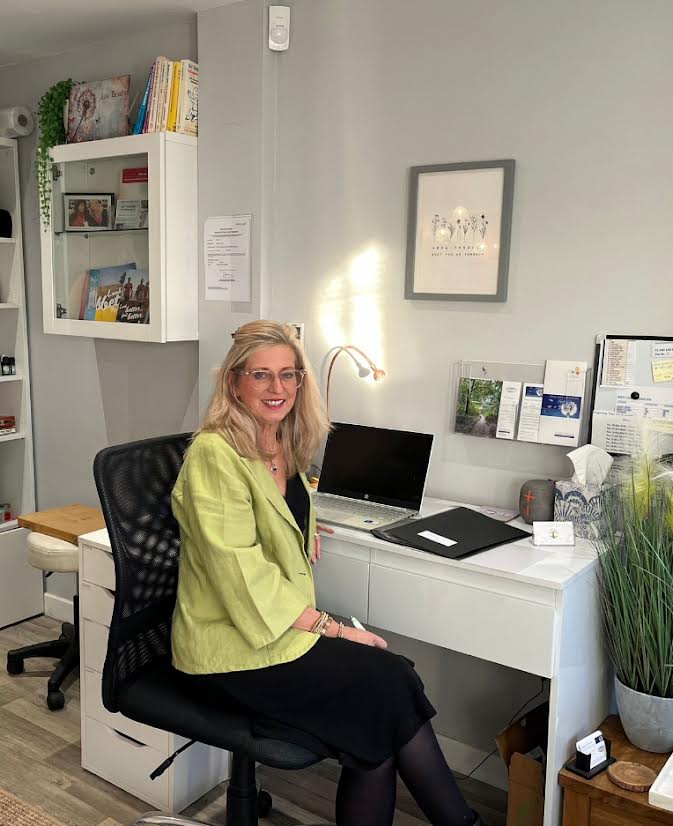 Carrie Vogel-Dennett, a professional coach, smiling warmly in a softly lit, modern office, conveying approachability and trust. She is seated, hands gently folded, with a calm, supportive demeanor. The background is blurred, focusing attention on her expression and presence.