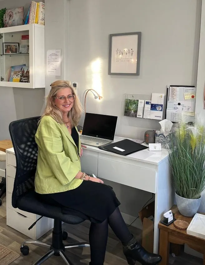 Carrie Vogel-Dennett, a professional coach, smiling warmly in a softly lit, modern office, conveying approachability and trust. She is seated, hands gently folded, with a calm, supportive demeanor. The background is blurred, focusing attention on her expression and presence.