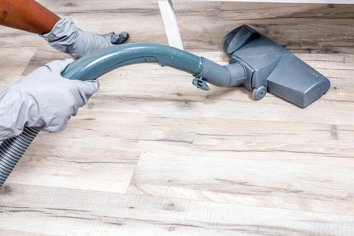 Hands in gloves holding a vacuum cleaner hose on a wooden floor.