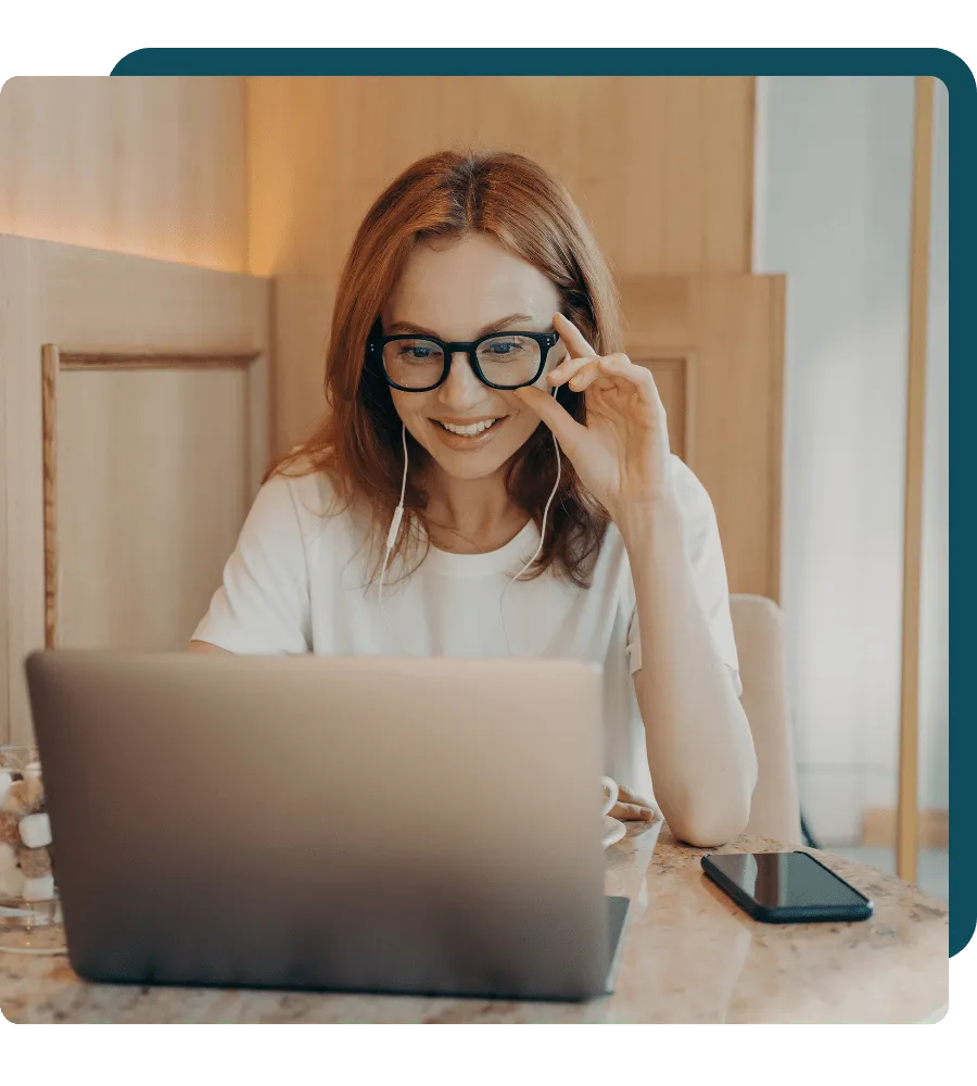 A woman smiling while adjusting her glasses and using a laptop with earphones, a smartphone, and a glass of water on the table.