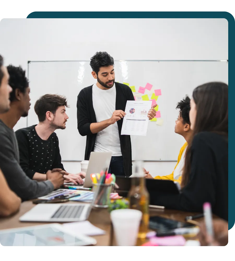 A man presenting a report with charts to a team during a business meeting, with laptops and coffee cups on the table.