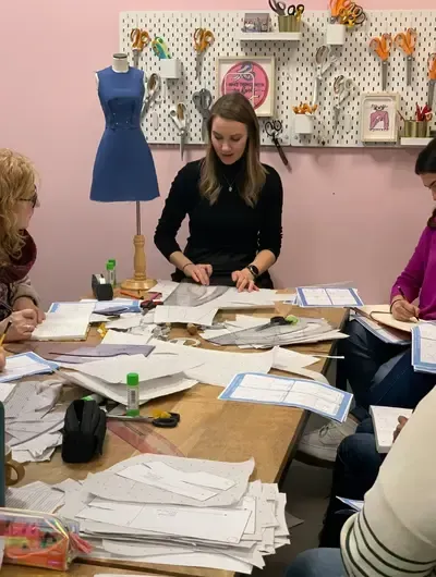 A sewing pattern workshop in progress, with an instructor guiding students around a table covered in paper pattern pieces, rulers, and glue sticks. A blue dress form and wall of sewing scissors are visible in the background. Perfect image for a fashion design or dressmaking class webpage.