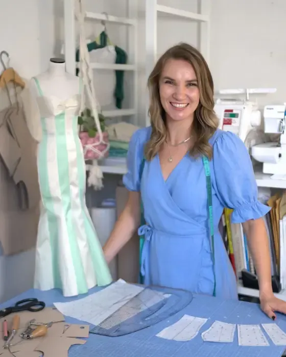 Smiling dressmaking instructor in a blue wrap dress standing beside a mannequin wearing a mint and cream striped gown. The sewing studio features pattern pieces, rulers, and a sewing machine, showcasing fashion design and garment construction in progress. Ideal image for a sewing course or dressmaking workshop webpage.