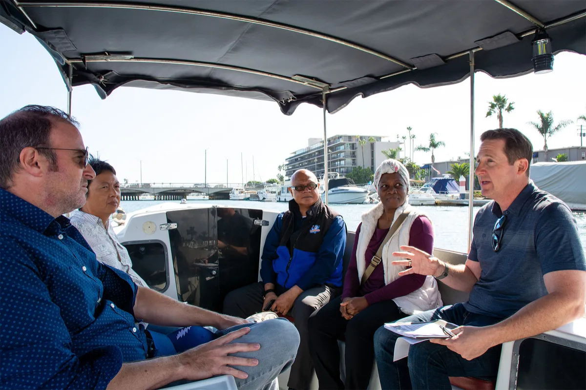 Group of four people conversing on a boat shaded by a canopy, with a harbor and buildings in the background.