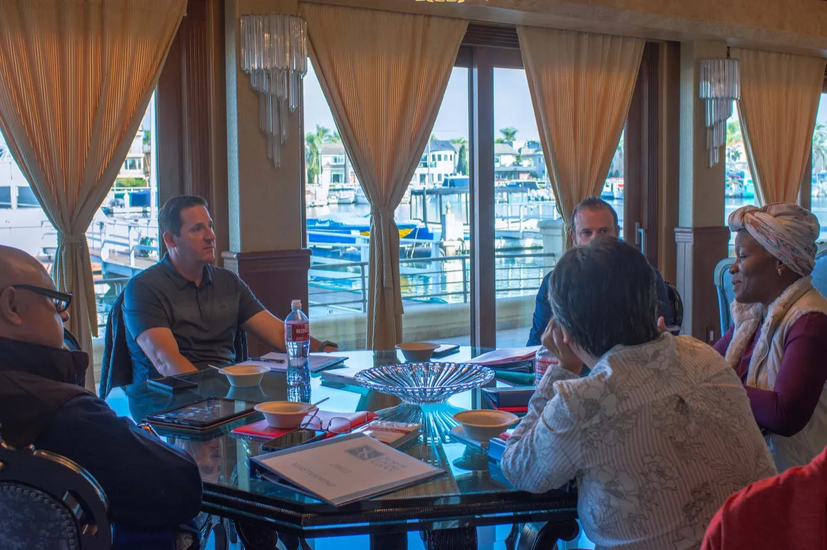 Five adults sitting in ornate black chairs around a dark polished dining table in a well-lit room with large windows, yellow curtains, and a crystal chandelier. The group appears to be in discussion.
