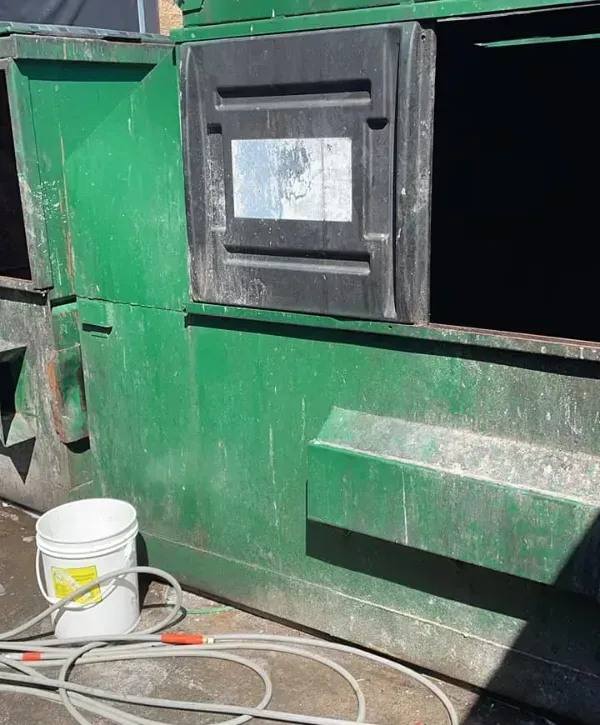 Worn green dumpster ready for pressure washing with hose and bucket nearby