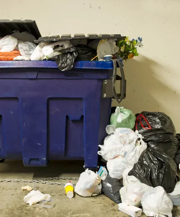 Overflowing blue commercial dumpster with trash bags and loose waste