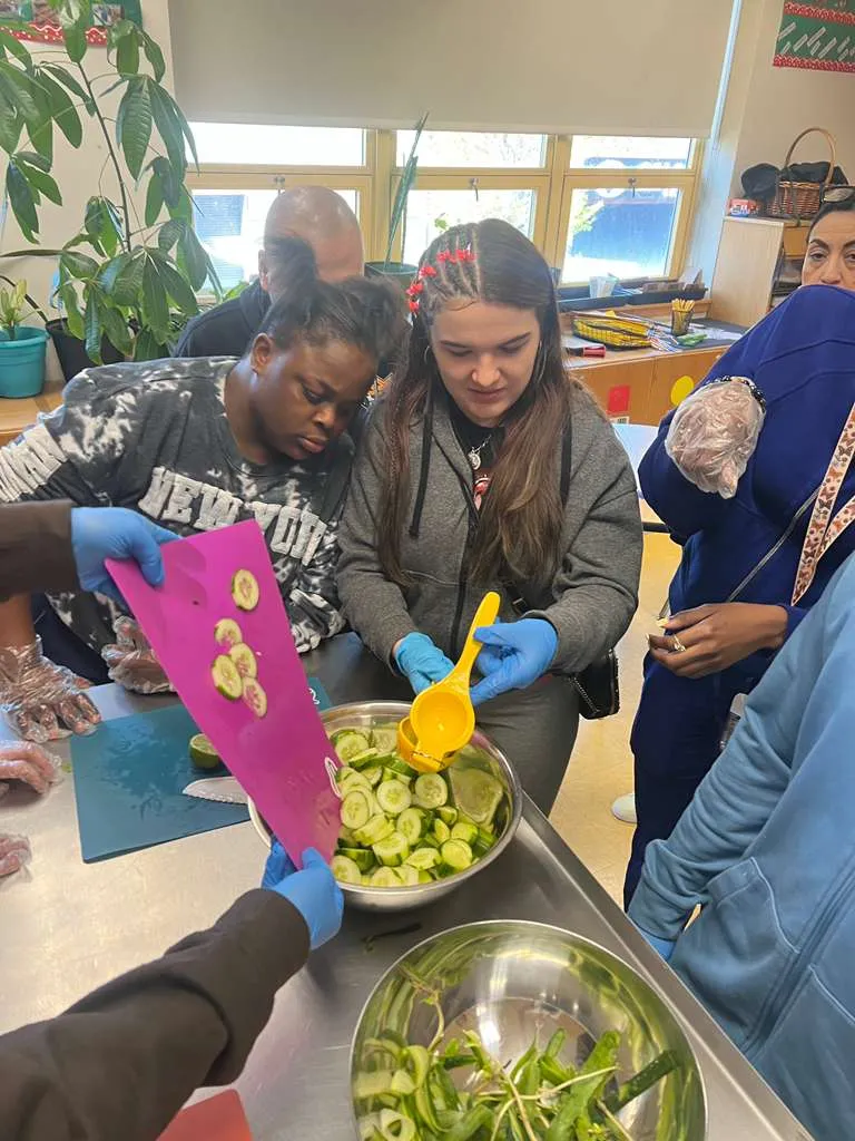 Students wearing gloves scooping sliced cucumbers from a mixing bowl during a cooking lesson