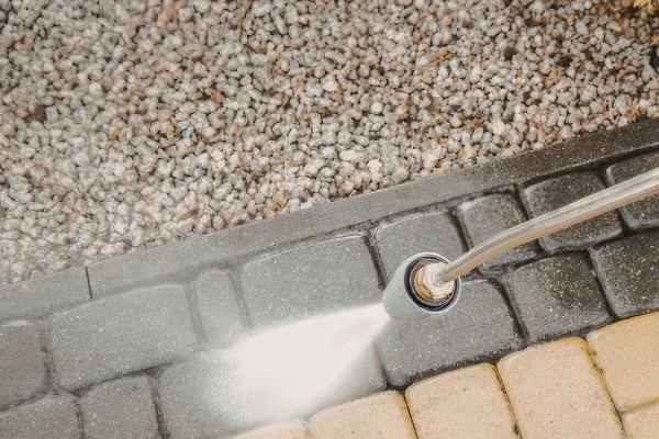 Overhead view of a pressure washer cleaning dirty grey and yellow patio pavers next to white gravel.