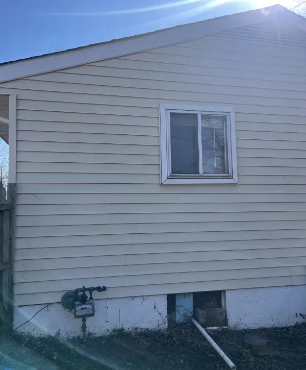 A cream-colored house exterior with a small window, gas meter, and open foundation access on a sunny day.