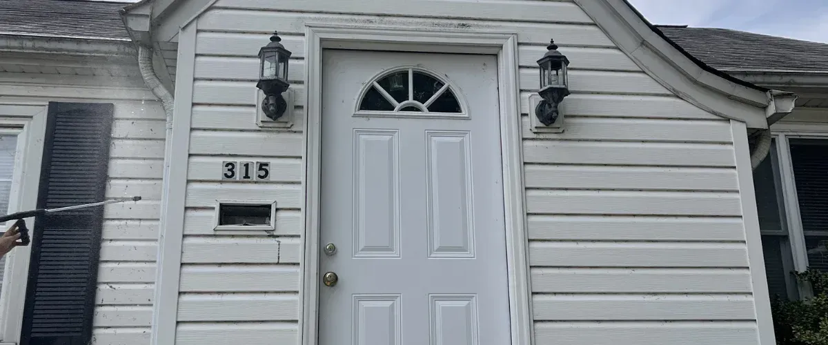 A house exterior with white siding and black shutters being pressure washed, showing dirt being removed.