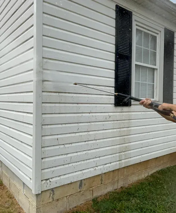 A person pressure washing dirty white vinyl house siding with a dark window and black shutters.
