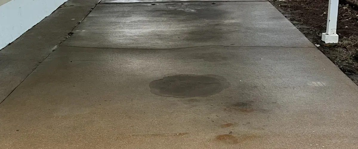 A damp concrete sidewalk with several puddles and darker stained areas, next to a white wall and some dirt.