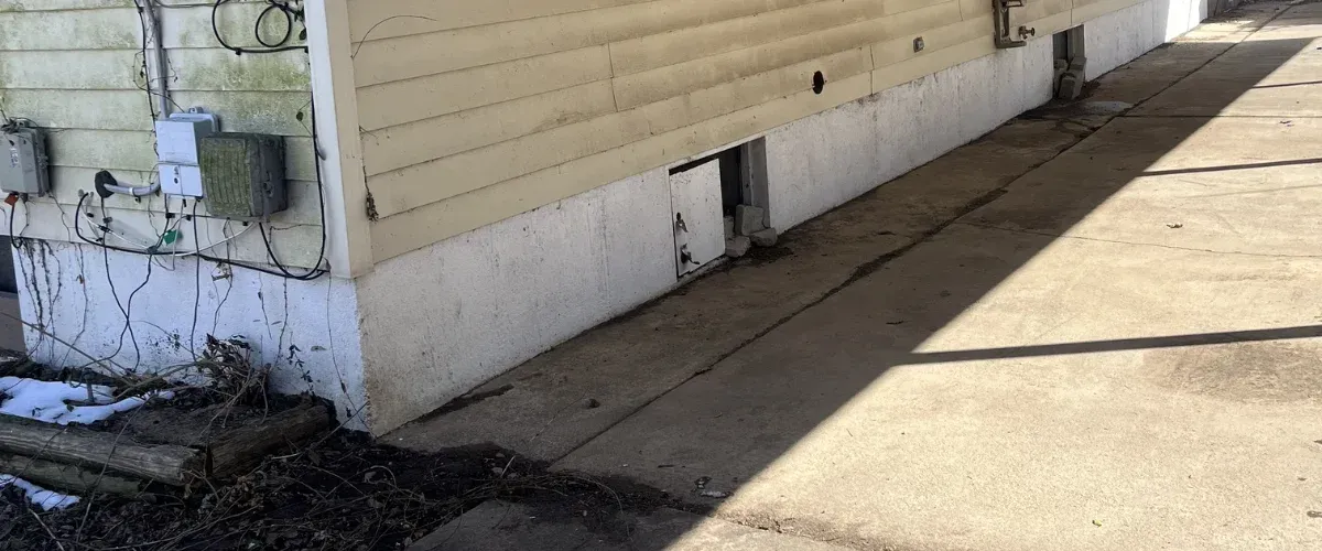 The foundation and lower exterior of a house with light yellow siding, concrete foundation, and a small open crawl space door.