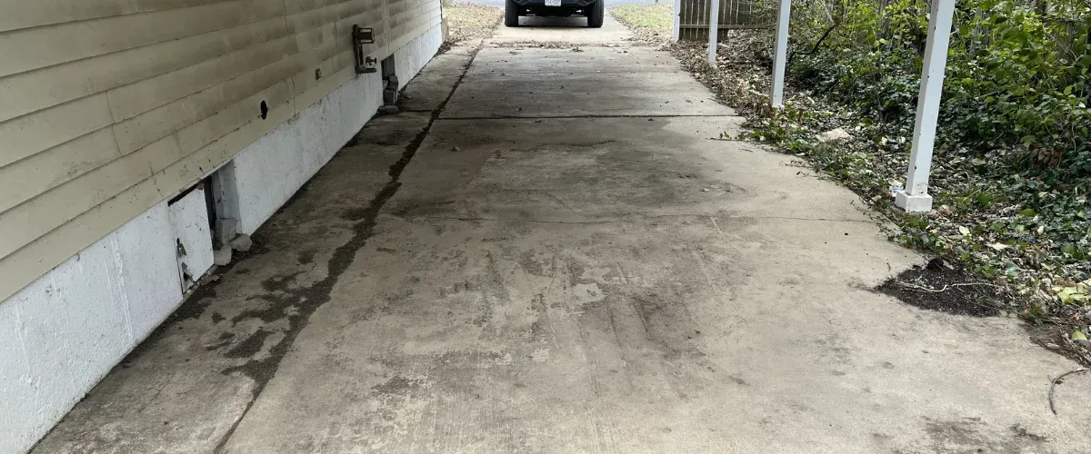 A long, narrow concrete driveway next to the side of a house with light yellow siding and a car parked in the distance.