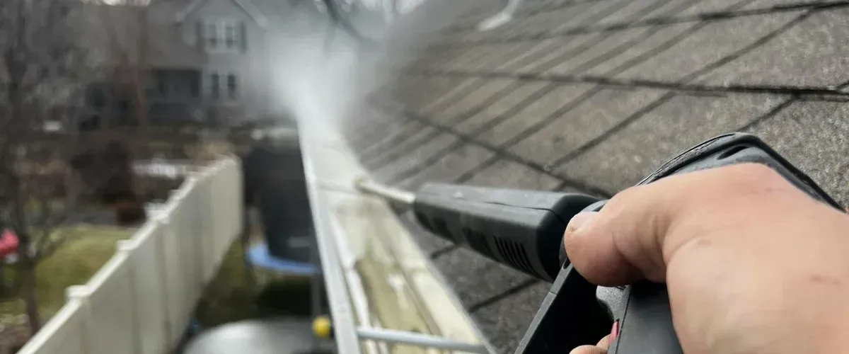 A person in blue jeans and black rubber boots powerwashing dirty patio slabs, showing clean and dirty sections.
