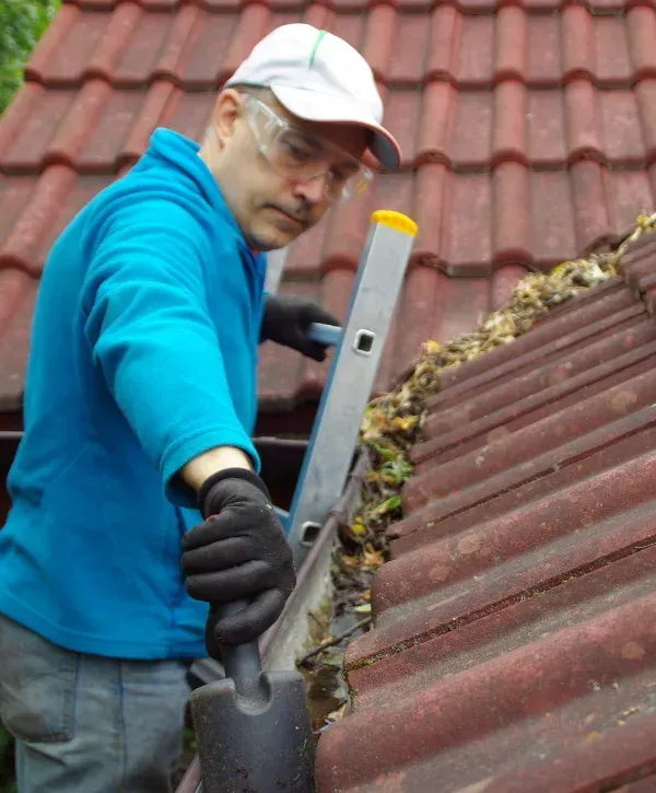 A man in safety glasses and gloves cleaning a rain gutter filled with leaves on a red tiled roof.