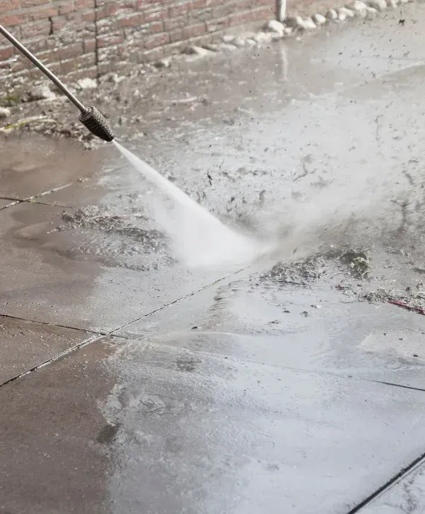A person in blue jeans and black rubber boots powerwashing dirty patio slabs, showing clean and dirty sections.