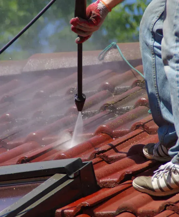A gloved hand uses a pressure washer to clean red roof tiles, seen from a low angle with a person's legs in jeans.