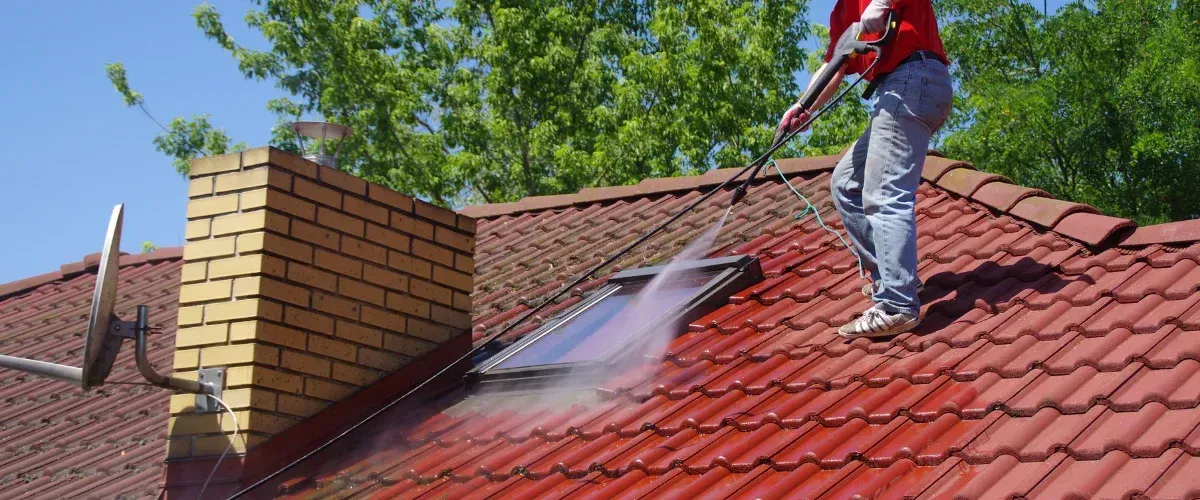 A man in jeans and a red shirt power washes a red tile roof with a skylight and brick chimney.