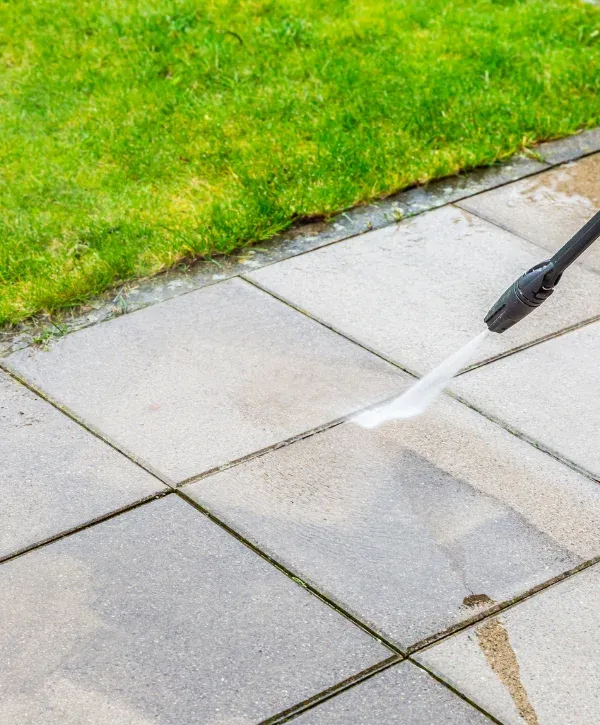 A close-up view of a patio being power-washed, with a stream of water cleaning the grey tiles next to green grass.