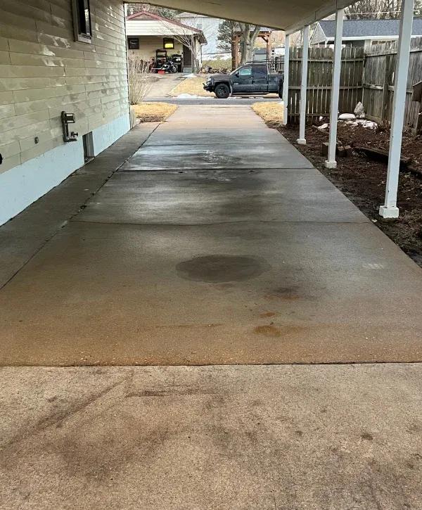 A long, damp concrete driveway extending from under a carport toward a black pickup truck in a suburban setting.