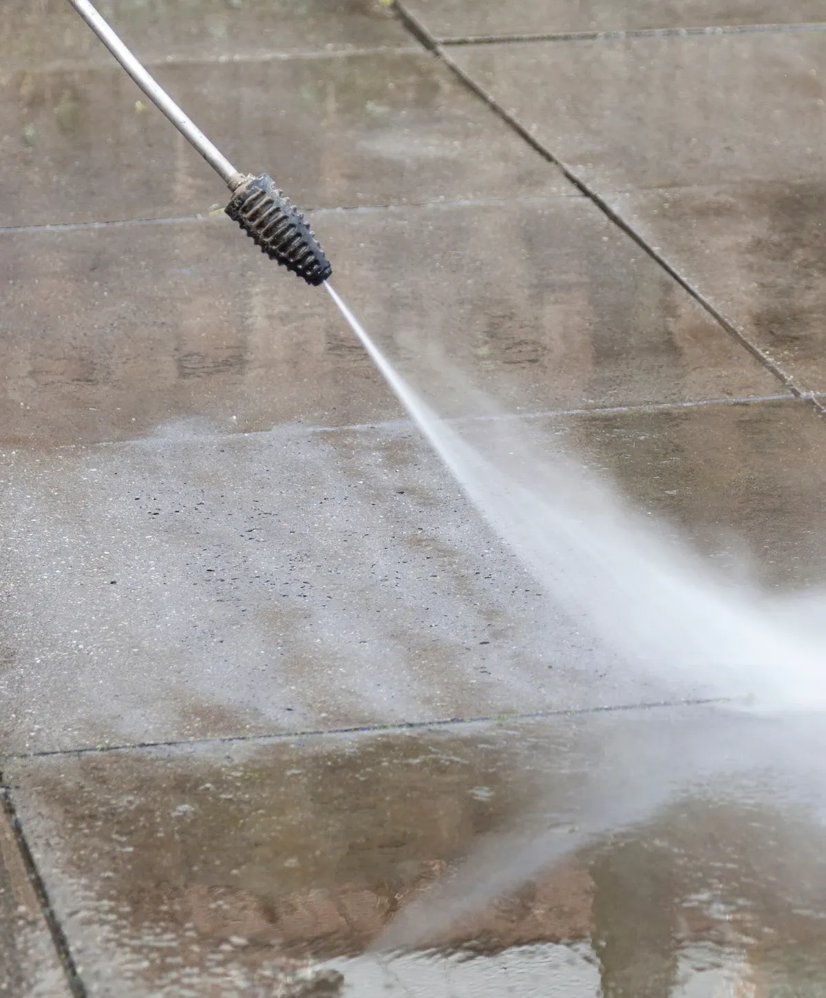 A pressure washer nozzle sprays a powerful jet of water onto a dirty paved patio, cleaning the surface.