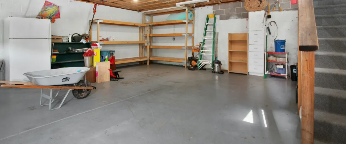 A wide shot of a cluttered garage or unfinished basement with a concrete floor, white refrigerator, wooden shelving, and a carpeted staircase.