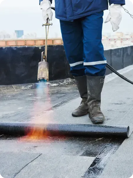 A person in work clothes and boots uses a torch to melt asphalt flat roofing material on a rooftop in Ottawa.