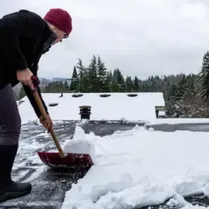 A worker is clearing snow off a flat roof, with snow-covered trees visible in the background under a cloudy sky.