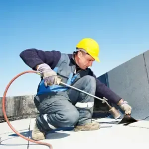 A roofer in a yellow hard hat uses a torch to seal the roof membrane during a flat roof repair under a clear blue sky.