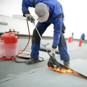 A roofer in blue overalls uses a blowtorch to install roofing material on a flat roof, with a red gas tank beside him.