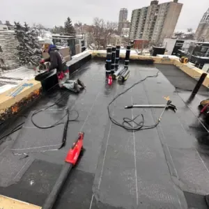 A crew is installing a new flat roof during winter, with snow still visible outside the building.