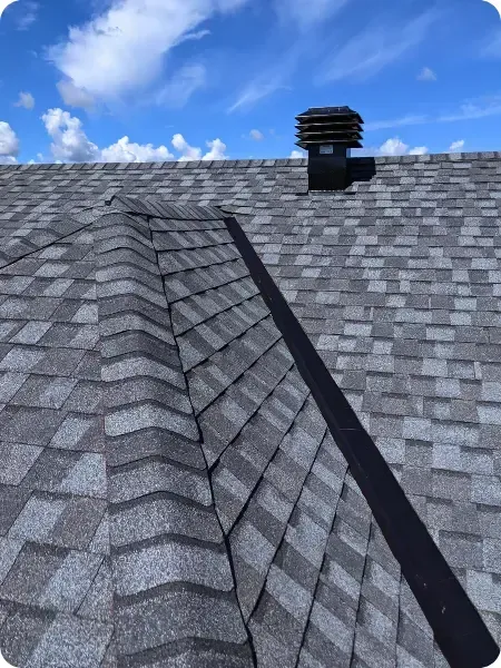 A close-up view of a grey shingled roof installed in Ottawa with a black vent pipe on the ridge under a bright blue sky with white clouds.