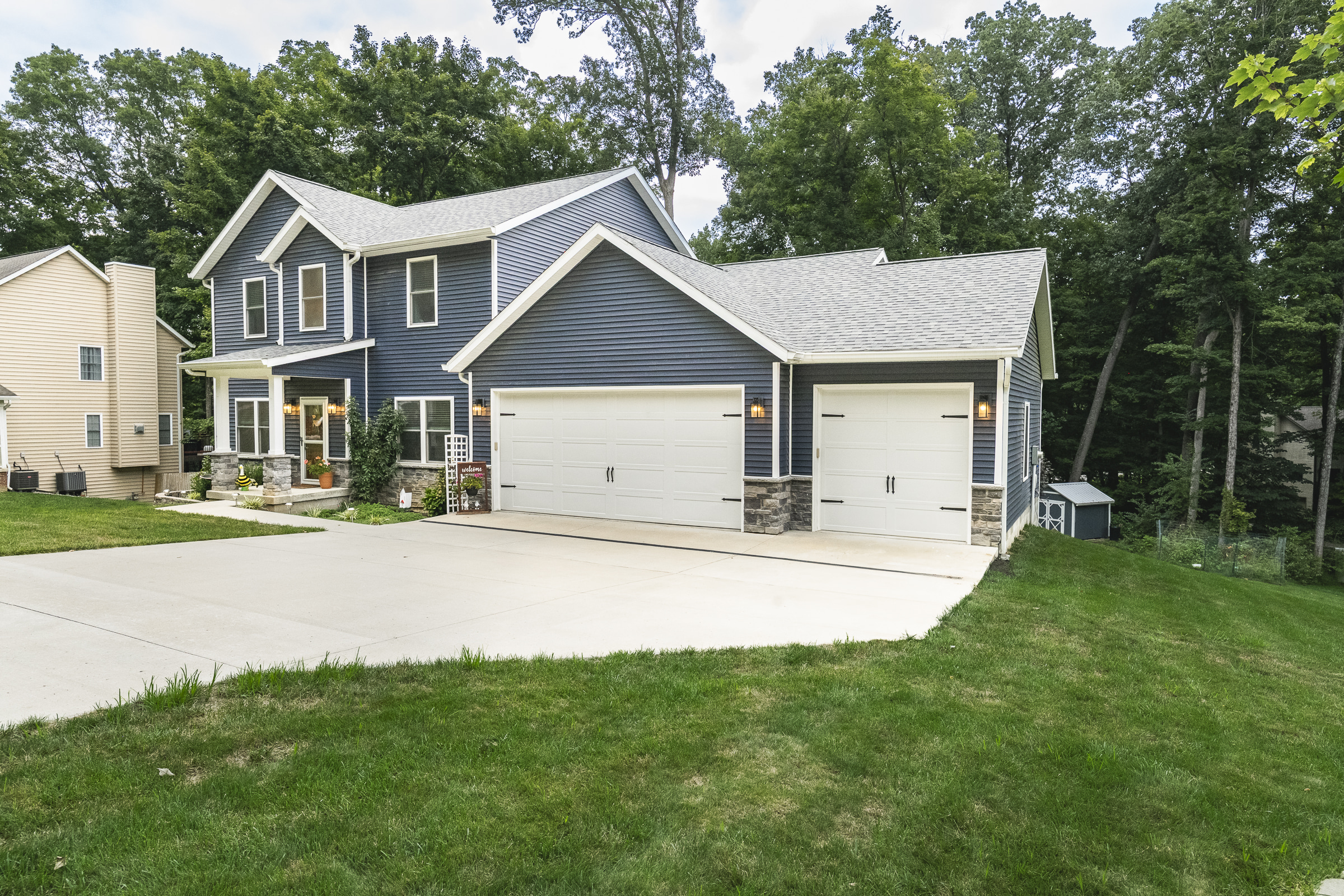 Blue two-story house with a three-car garage, concrete driveway, and wooded backyard in Ellettsville, Indiana