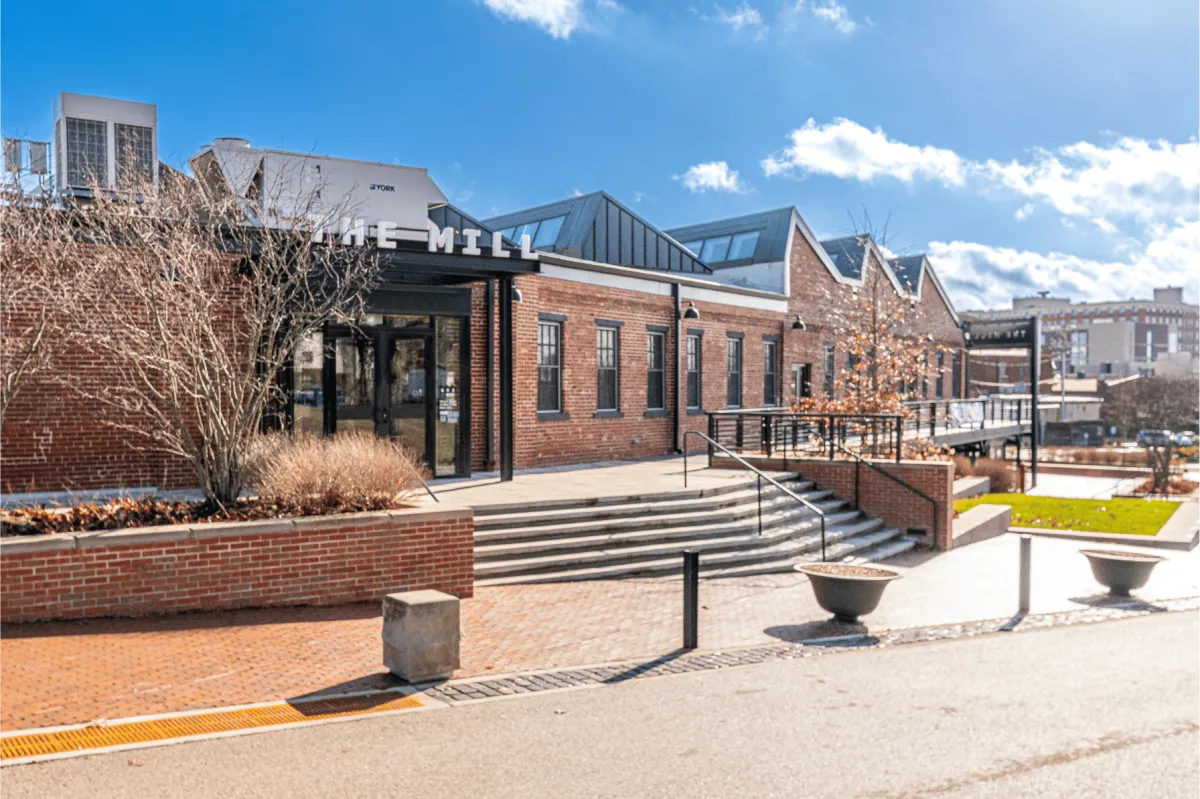 Exterior of The Mill, a brick coworking and entrepreneurship hub in downtown Bloomington, Indiana, shown on a sunny day with steps, railing, and blue sky.