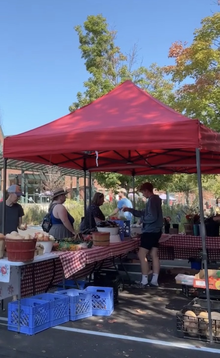 Vendor booth at the Bloomington Community Farmers’ Market with shoppers browsing fresh produce under a red canopy on a sunny day.