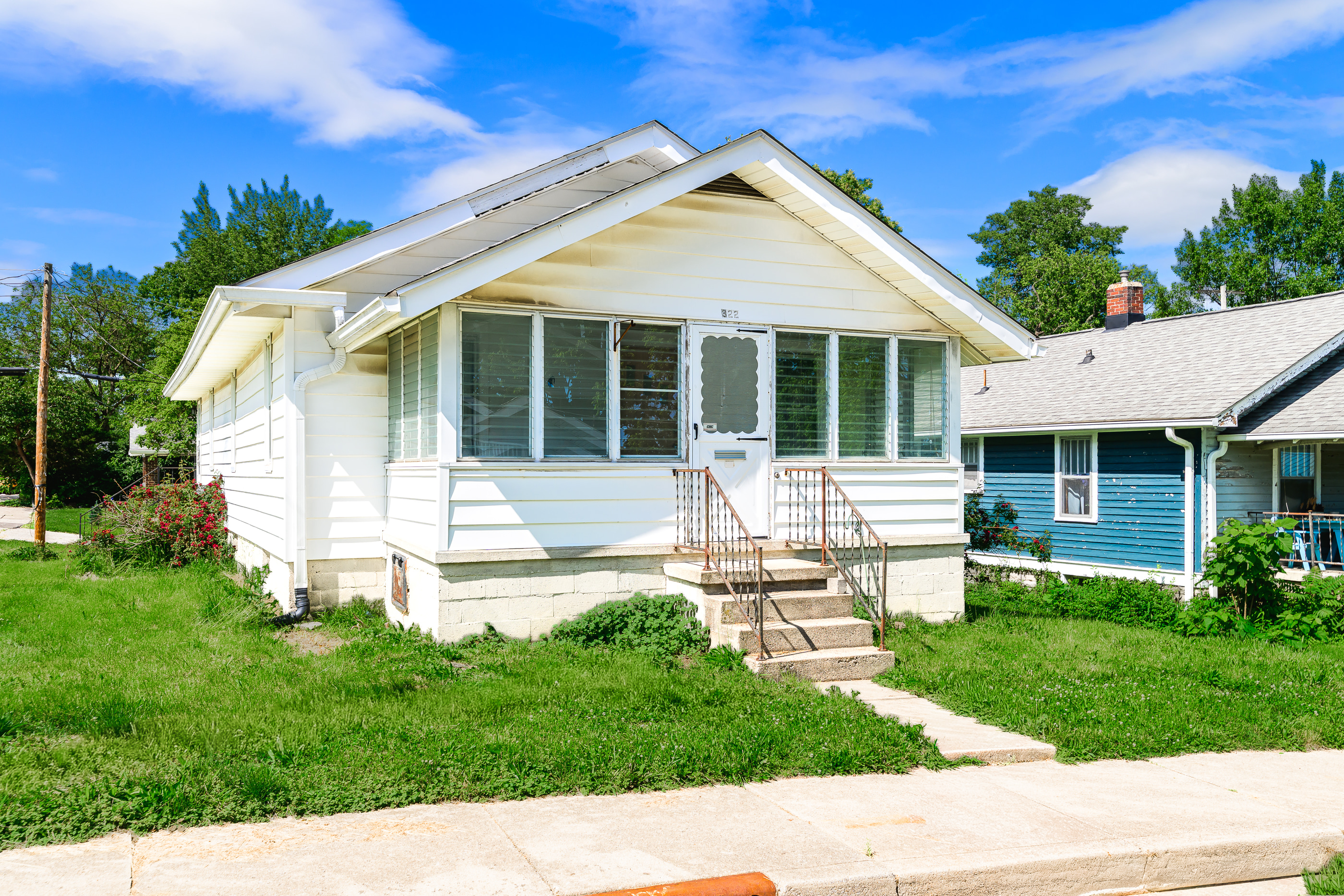Small white bungalow-style home with enclosed front porch, steps, green lawn, and blue sky in a residential neighborhood.