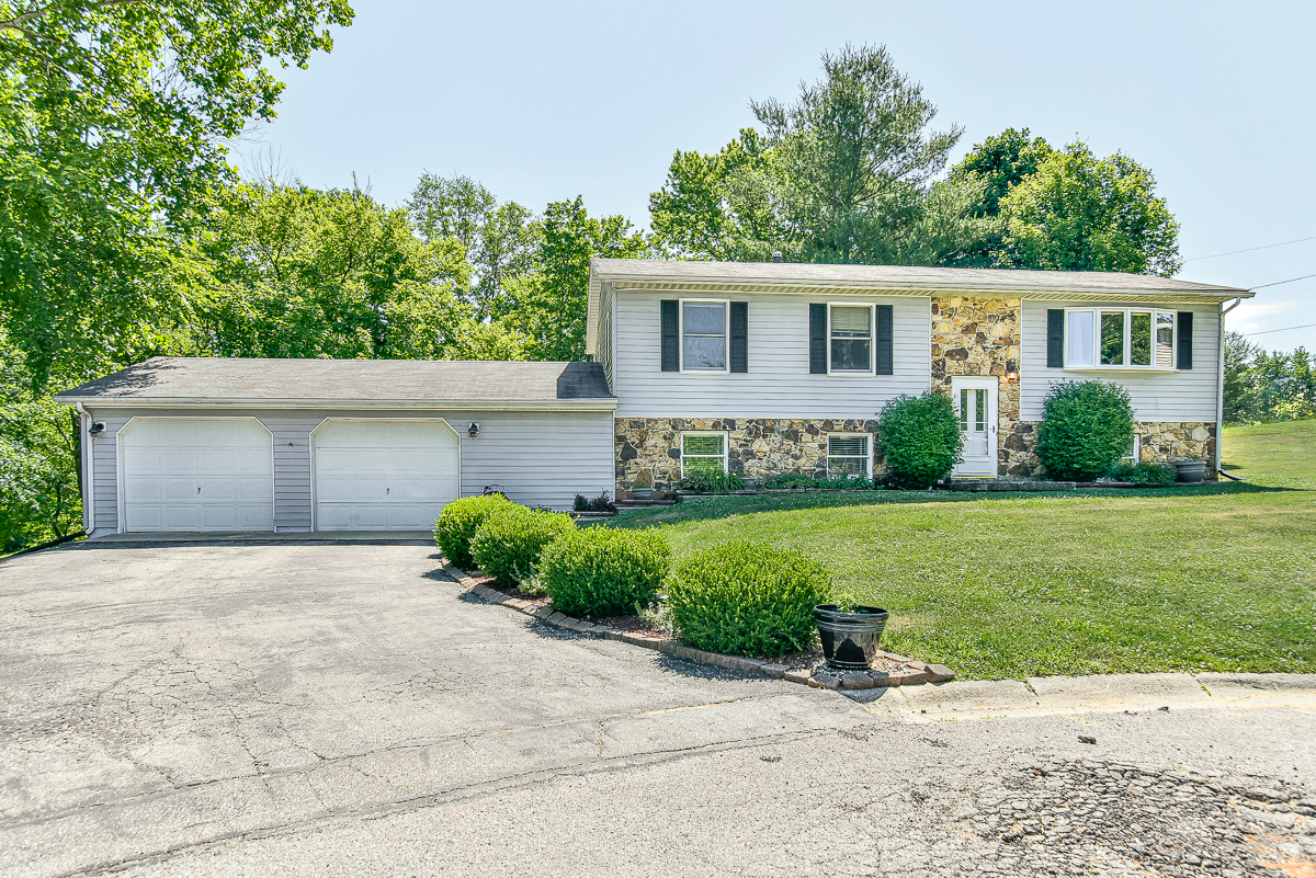 Split-level home with light gray siding and stone accent facade, attached two-car garage, and landscaped front yard along a paved driveway.