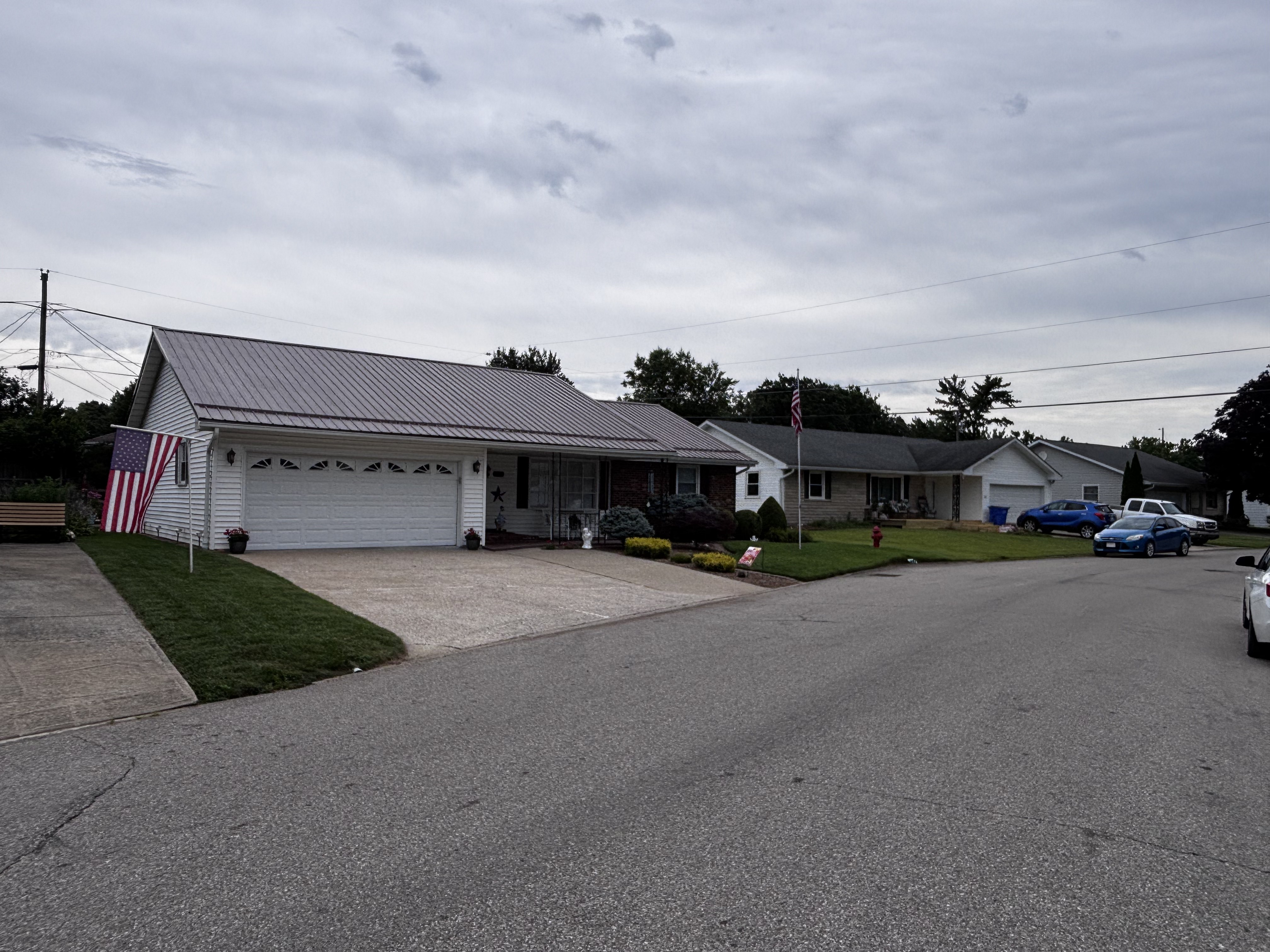 White ranch-style home with an attached two-car garage, standing-seam metal roof, brick accents, and an American flag, sitting on a corner lot in south-central Indiana