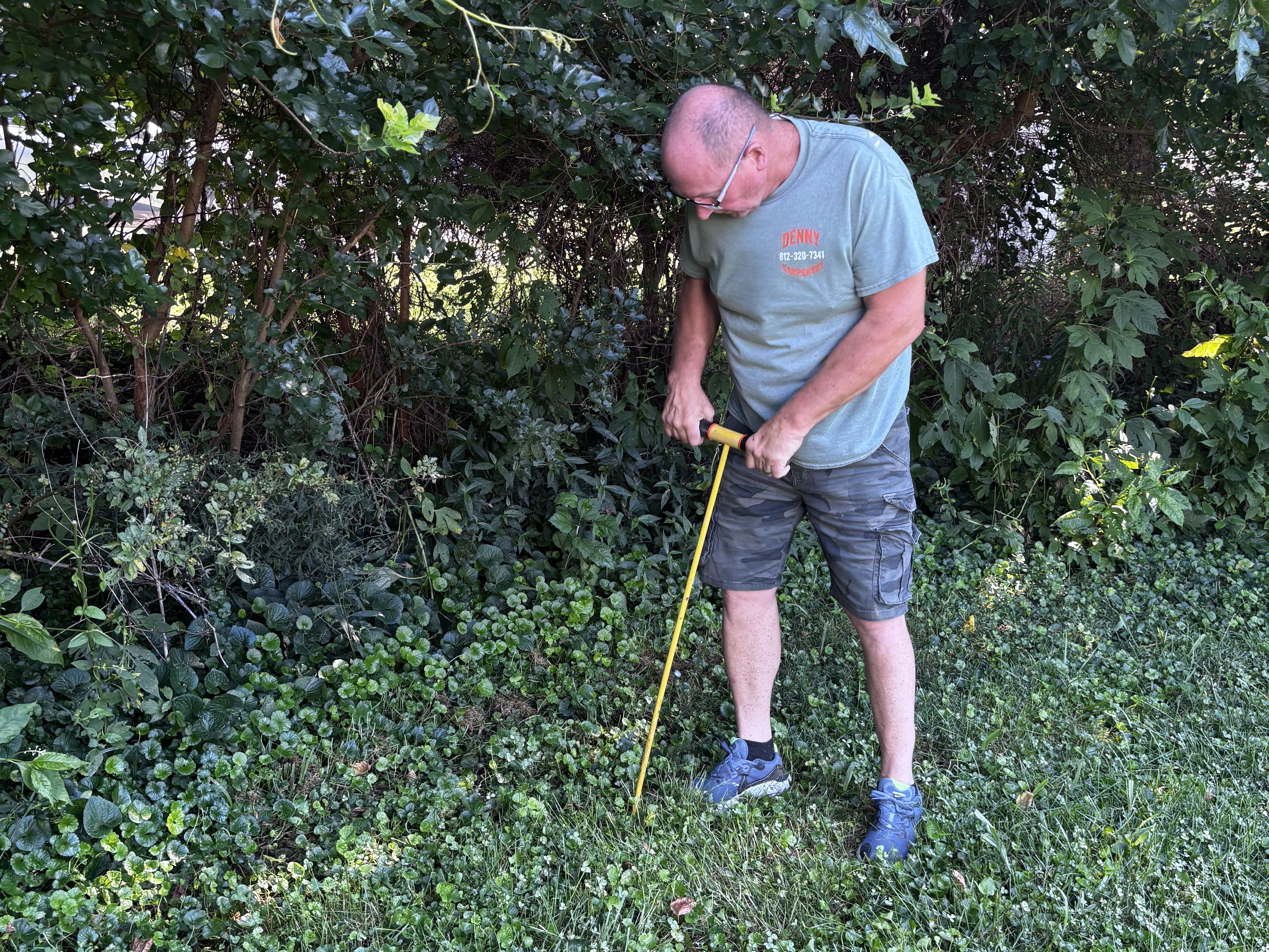 Technician using a metal probe rod to locate a septic tank in an overgrown yard before inspection