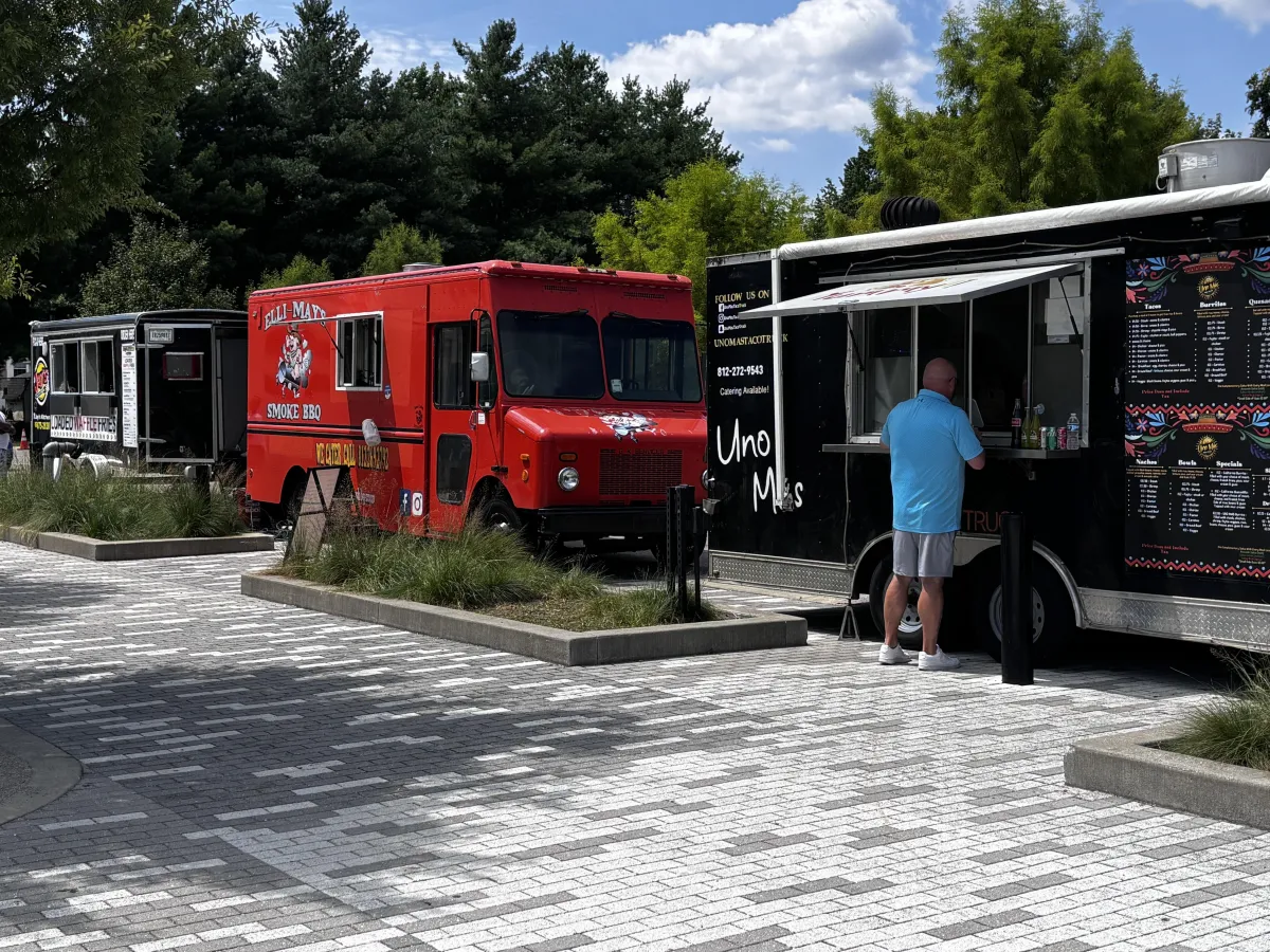Food trucks, including Elli-May's Smoke BBQ and a taco truck, parked at Switchyard Park in Bloomington, Indiana, with a customer ordering at the window on a sunny afternoon